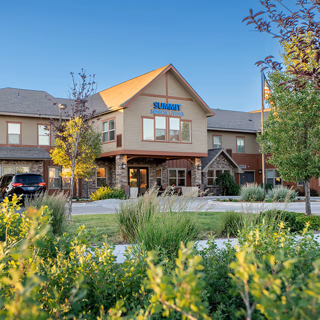 Exterior view of Summit Senior Living facility with a two-story building featuring a peaked roof, stone and wood siding, surrounded by trees, shrubs, and a well-maintained lawn under a clear blue sky.