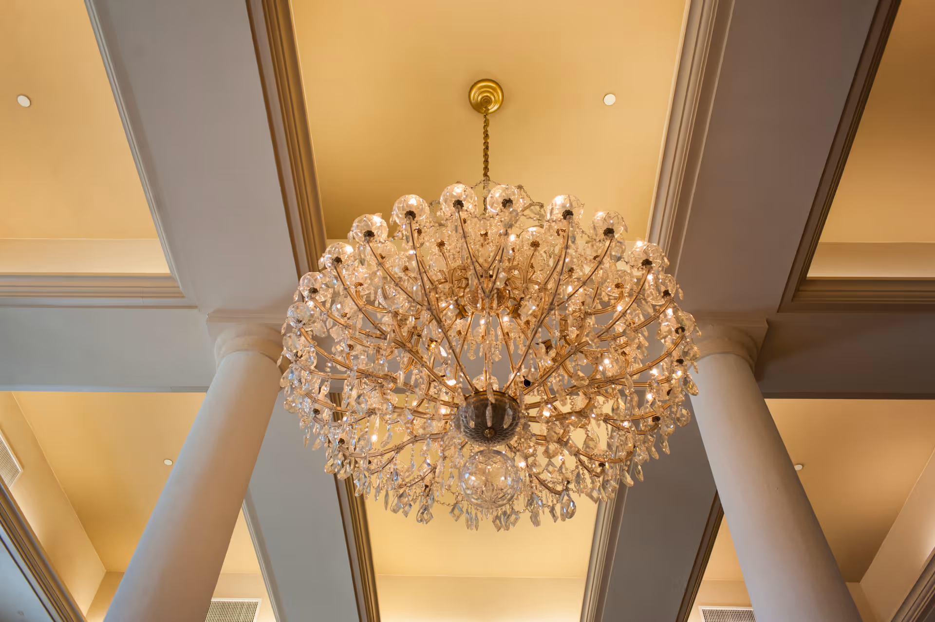 A large, ornate crystal chandelier hanging from a high ceiling with decorative molding and columns in the background.