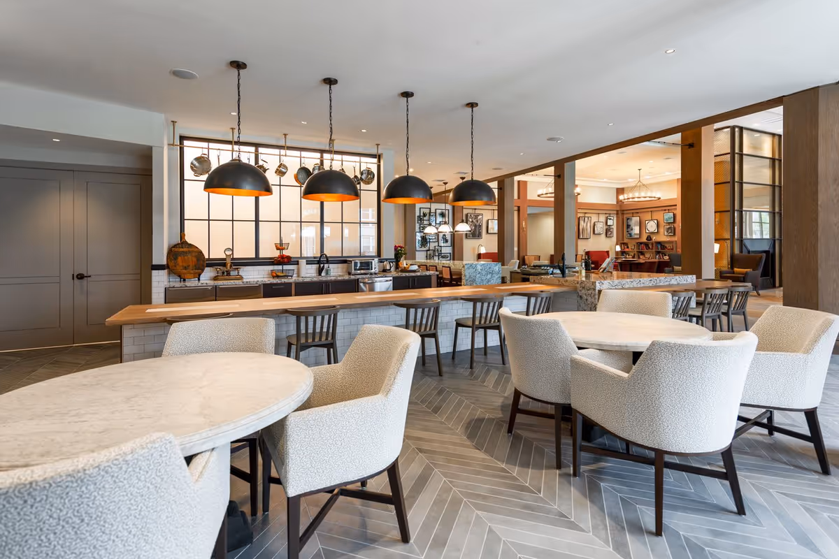 Open communal dining area with round marble tables, upholstered chairs, and a long island with pendant lights and a serving counter.