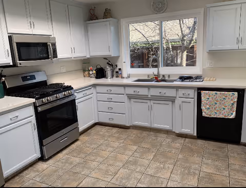 A clean and modern kitchen with white cabinets, a stainless steel gas stove and microwave, a black dishwasher with a colorful towel hanging on its handle, a double sink under a window showing a view of a tree and a fence outside, and a tiled floor.