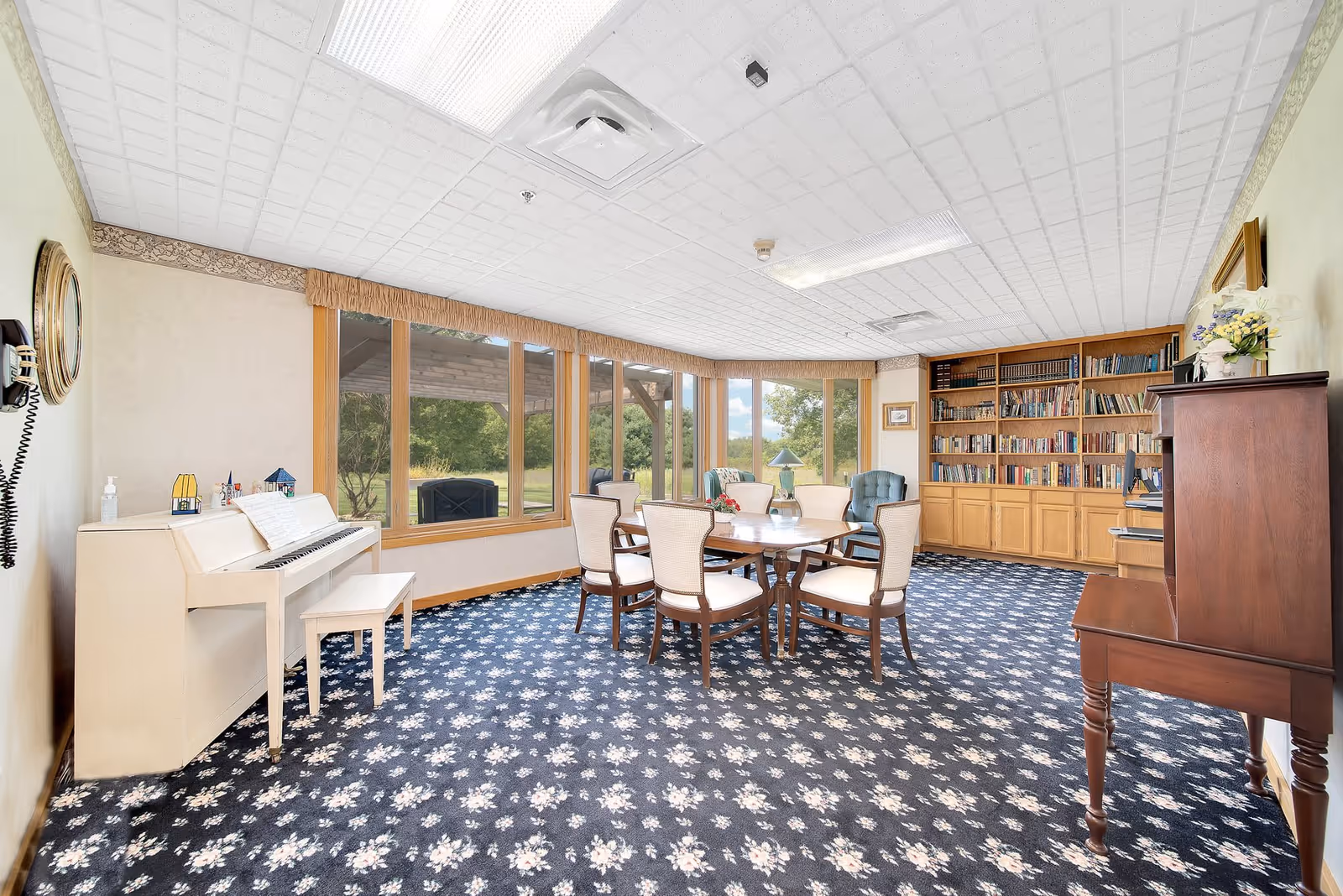 A bright and spacious common room with large windows overlooking greenery. The room features a round wooden table surrounded by six cushioned chairs, a white piano with a matching bench, a wooden bookshelf filled with books, a blue upholstered armchair, and a wooden cabinet with a vase of flowers on top. The floor is covered with a blue carpet patterned with white flowers, and the ceiling has white tiles with fluorescent lighting.