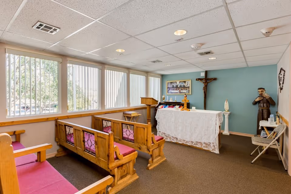 Small chapel room with wooden pews featuring red cushions, a white altar covered with a lace cloth, religious statues, a crucifix on the wall, and large windows with vertical blinds letting in natural light.