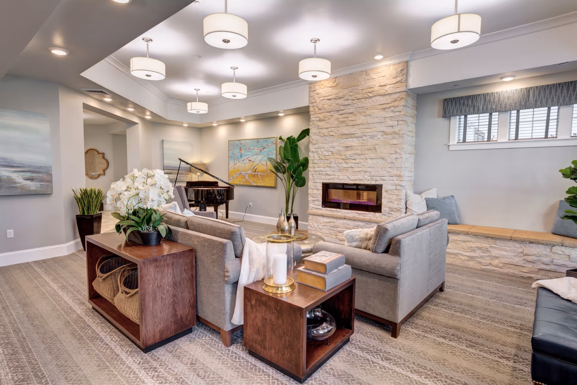 A modern living room with two gray sofas arranged around a stone fireplace. The room features a patterned carpet, a grand piano in the corner, several plants, and contemporary ceiling lights. There are paintings on the walls and a window with a cushioned bench underneath.