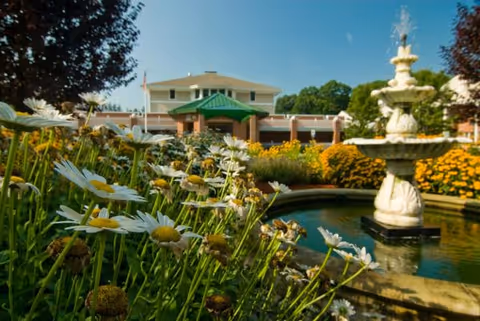 A garden area with blooming white daisies and yellow flowers surrounding a stone fountain with water spraying from the top. In the background, there is a two-story building with a green roof over the entrance, under a clear blue sky.