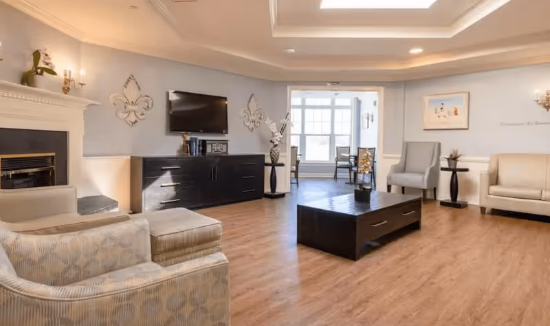 A spacious living room with light blue walls, wooden flooring, and a tray ceiling with recessed lighting. The room features a fireplace on the left, a wall-mounted flat-screen TV above a dark wooden cabinet, a patterned armchair with an ottoman, a gray armchair, a beige sofa, and a dark wooden coffee table in the center. Decorative wall accents and framed artwork are visible, along with a windowed dining area in the background.