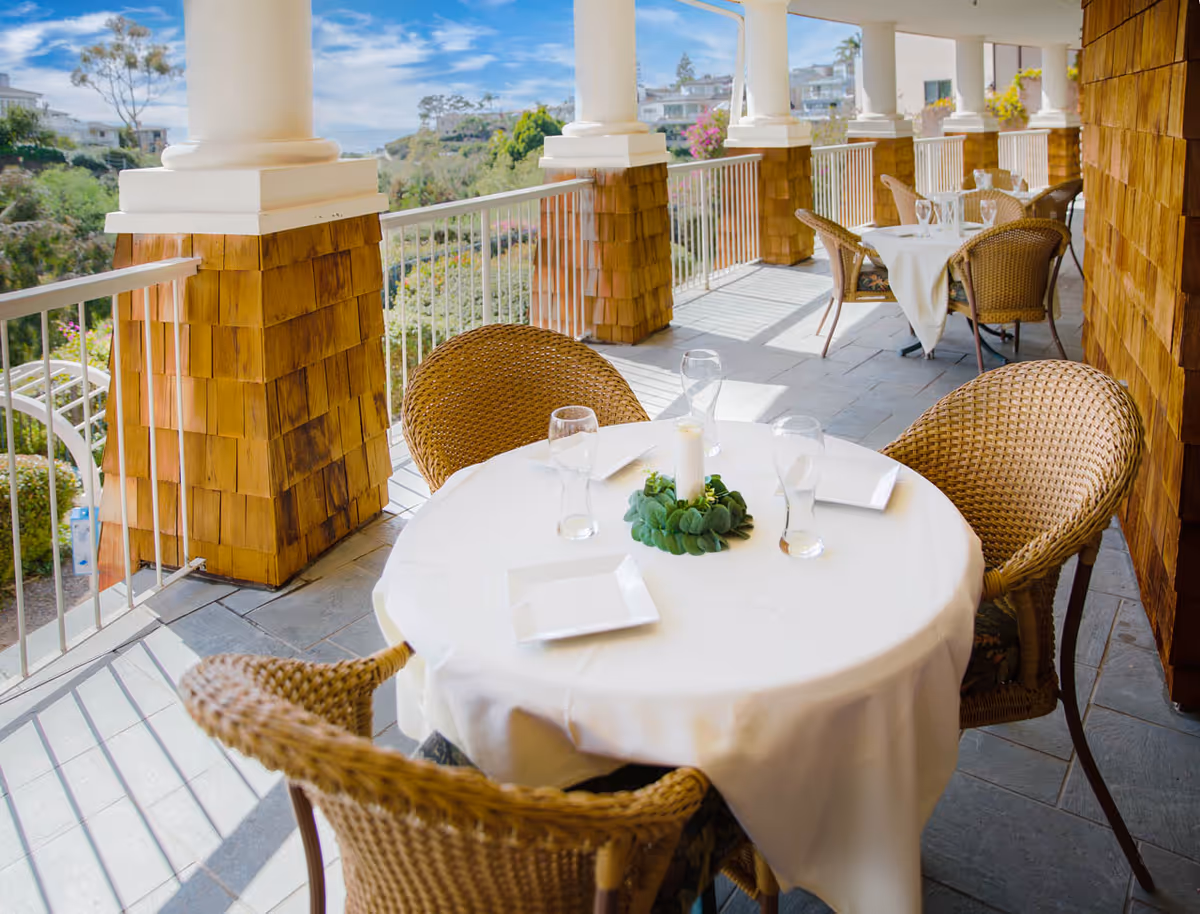 Outdoor covered patio area with round tables covered in white tablecloths, wicker chairs, and glassware. The patio overlooks a garden with greenery and houses in the distance under a partly cloudy sky.
