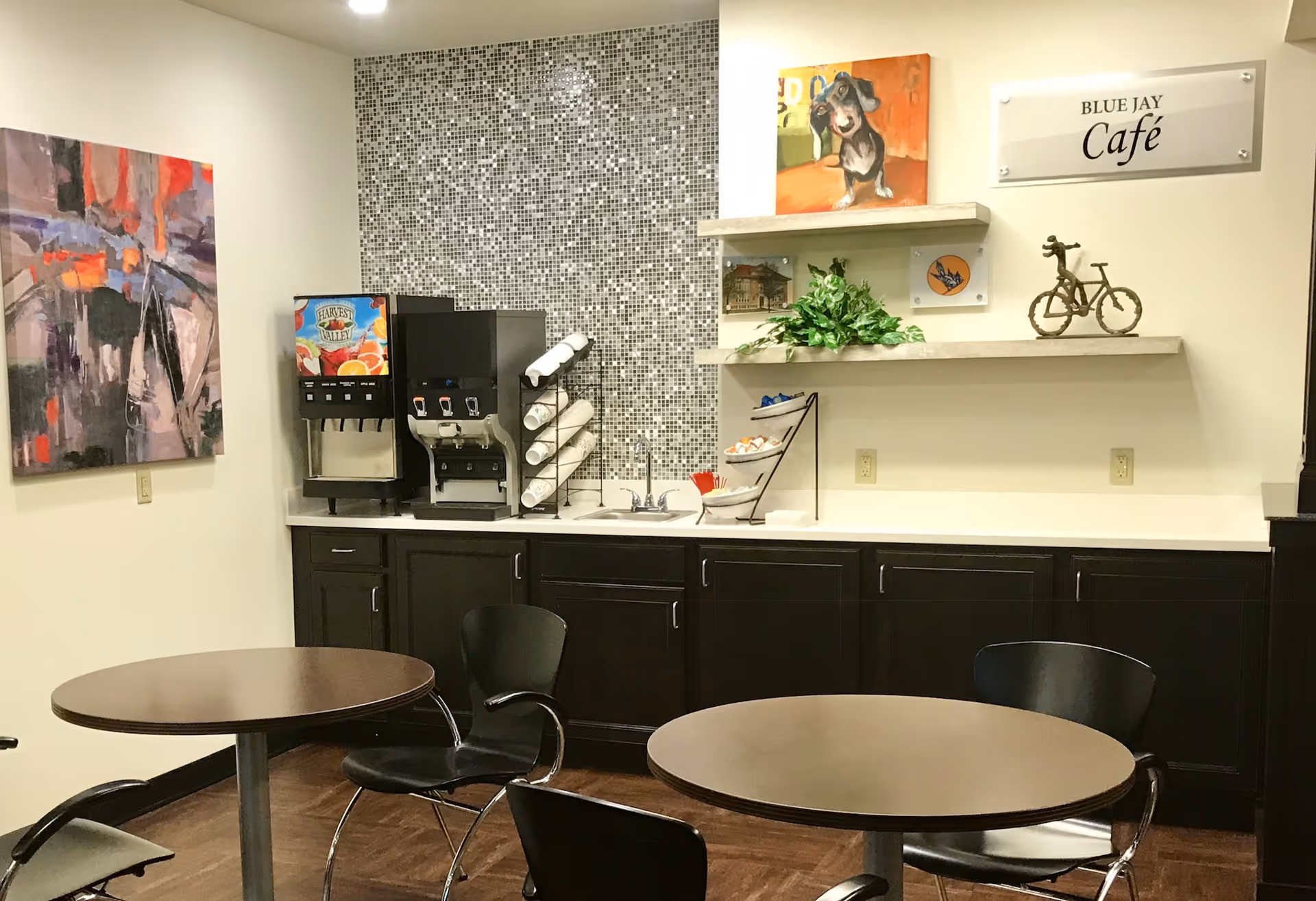 Interior view of a small café area named Blue Jay Café with two round tables and black chairs. The back wall features a beverage dispenser, a small sink, dark cabinets, and decorative shelves with a plant, a small bicycle sculpture, and artwork including a painting of a dog. The wall behind the beverage dispenser has a mosaic tile backsplash.