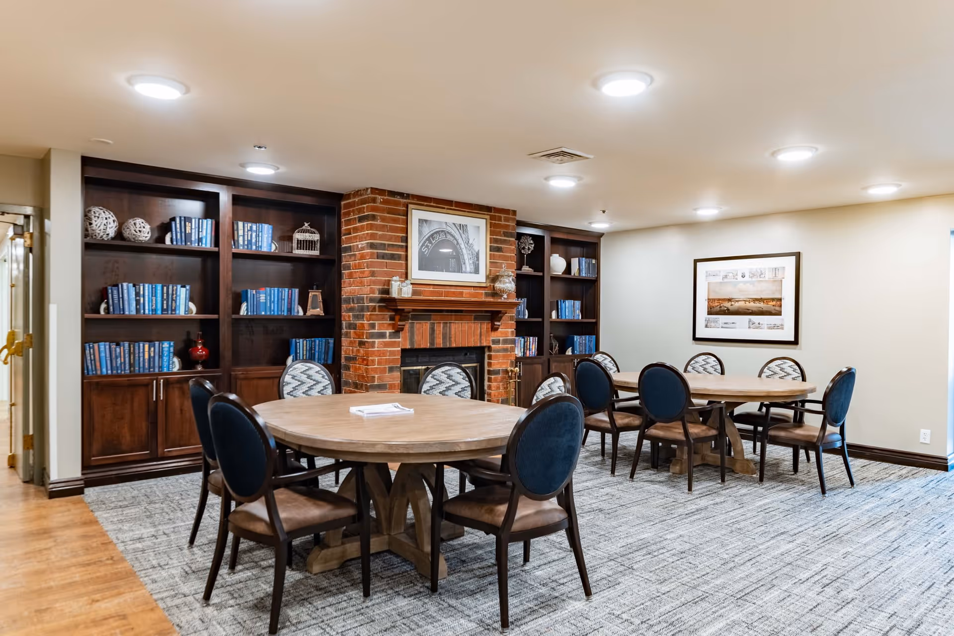 A cozy senior living common area with two round wooden tables surrounded by chairs with dark blue cushions. The room features a brick fireplace with a framed black and white photo above it, flanked by dark wooden bookshelves filled with blue books and decorative items. The floor is carpeted with a patterned rug, and the walls are painted light beige with a framed picture on one side.