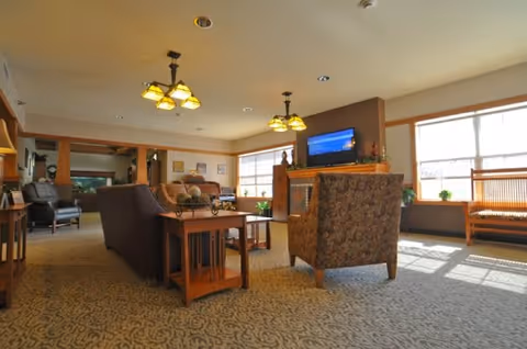 A spacious living room area with patterned carpet, multiple upholstered chairs and sofas, wooden side tables, large windows letting in natural light, a fireplace with a TV mounted above it, and ceiling lights with yellow shades.