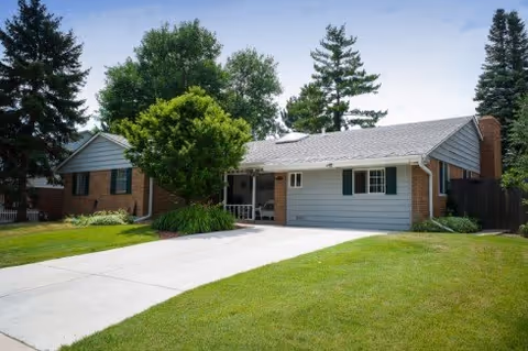 Single-story residential building with a gray roof, light gray siding, and brick accents. The house has green shutters on the windows and a driveway leading up to a covered porch area. The front yard is well-maintained with green grass, a tree, and shrubs. Tall trees are visible in the background under a partly cloudy sky.