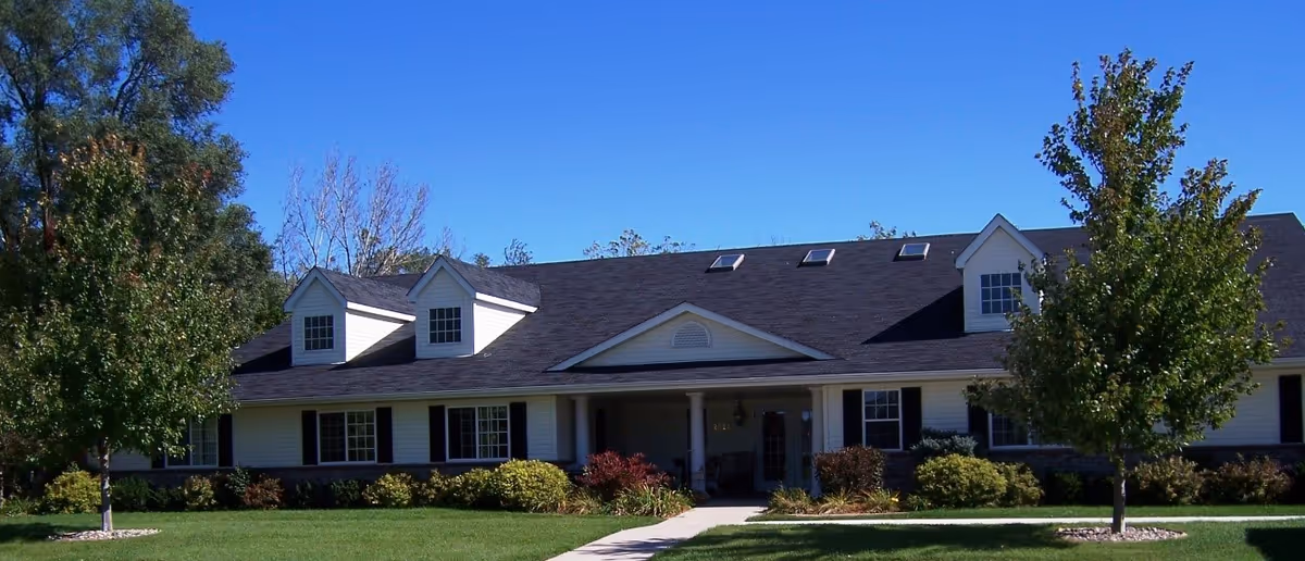 Exterior view of a single-story residential building with a dark shingled roof, white siding, and multiple windows. The building has a covered entrance with columns and is surrounded by green grass, bushes, and trees under a clear blue sky.