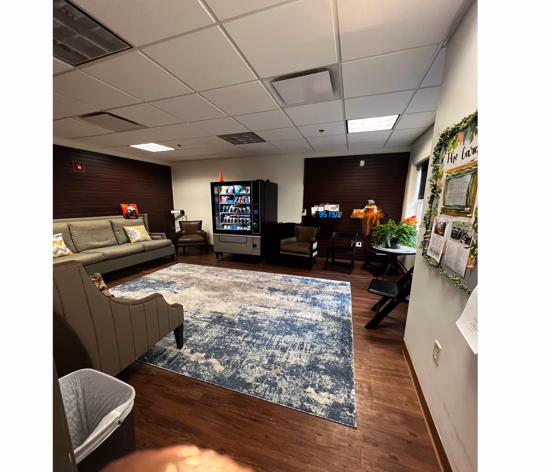 A cozy lounge area with a gray sofa, two brown armchairs, a vending machine stocked with snacks, a patterned blue and beige rug on wooden flooring, and a bulletin board decorated with greenery on the right wall.