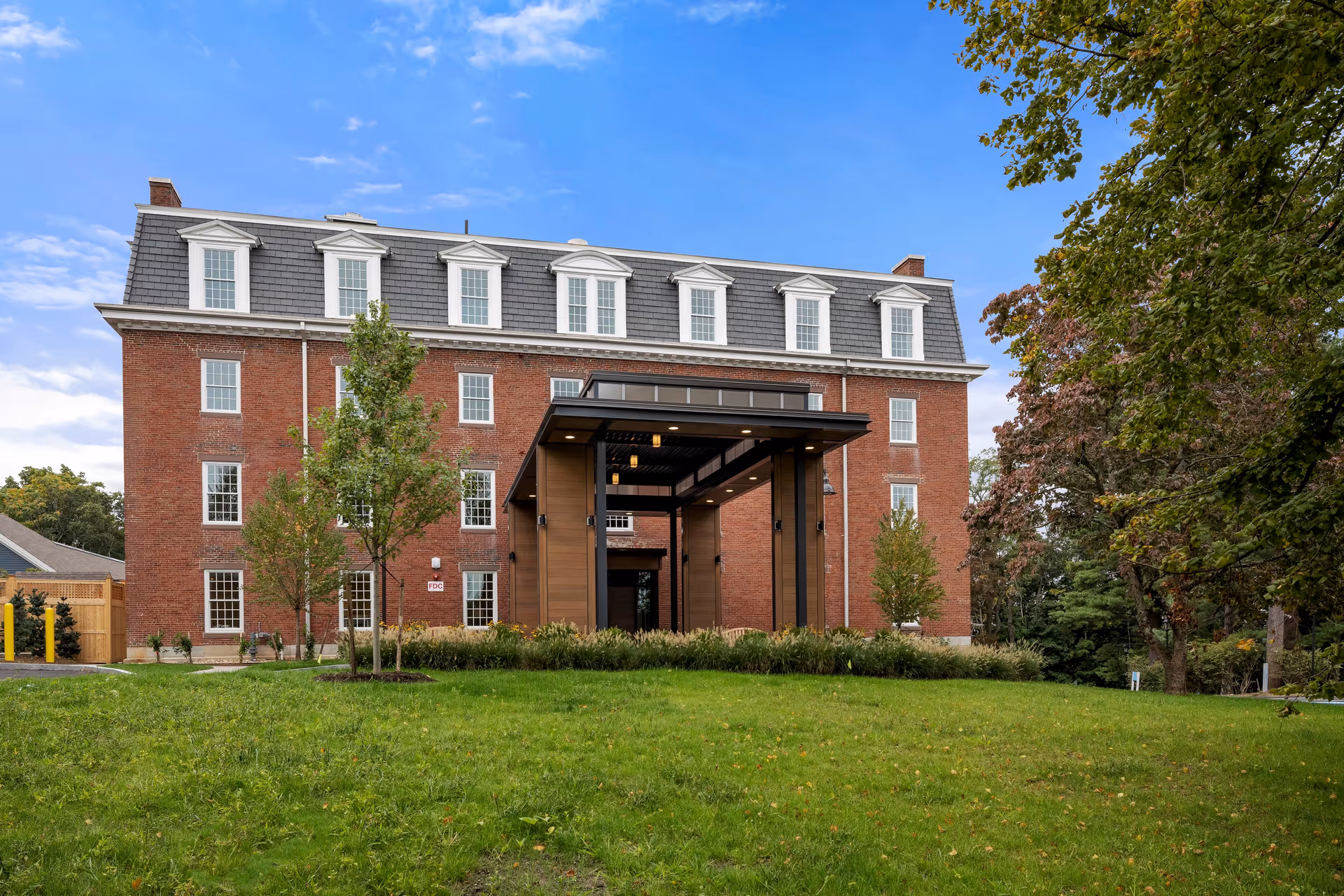 A large brick building with multiple windows and a gray mansard roof, featuring a covered entrance with wooden and black metal accents. The building is surrounded by green grass, small trees, and other foliage under a partly cloudy blue sky.