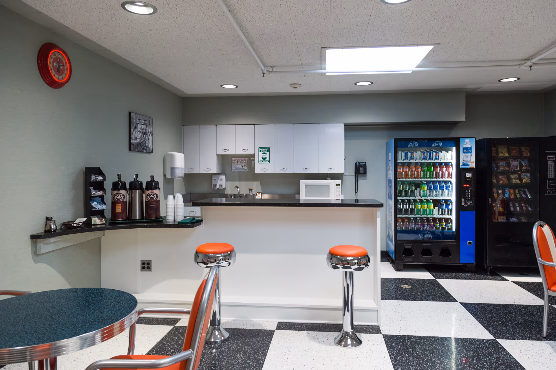 A small kitchenette area with a counter and two orange cushioned stools. Behind the counter are white cabinets, a microwave, a sink, and a wall-mounted phone. To the right, there are vending machines stocked with drinks and snacks. On the left side, there is a coffee station with coffee dispensers, cups, and condiments. The floor has a black and white checkered pattern, and there is a round table with orange chairs in the foreground.