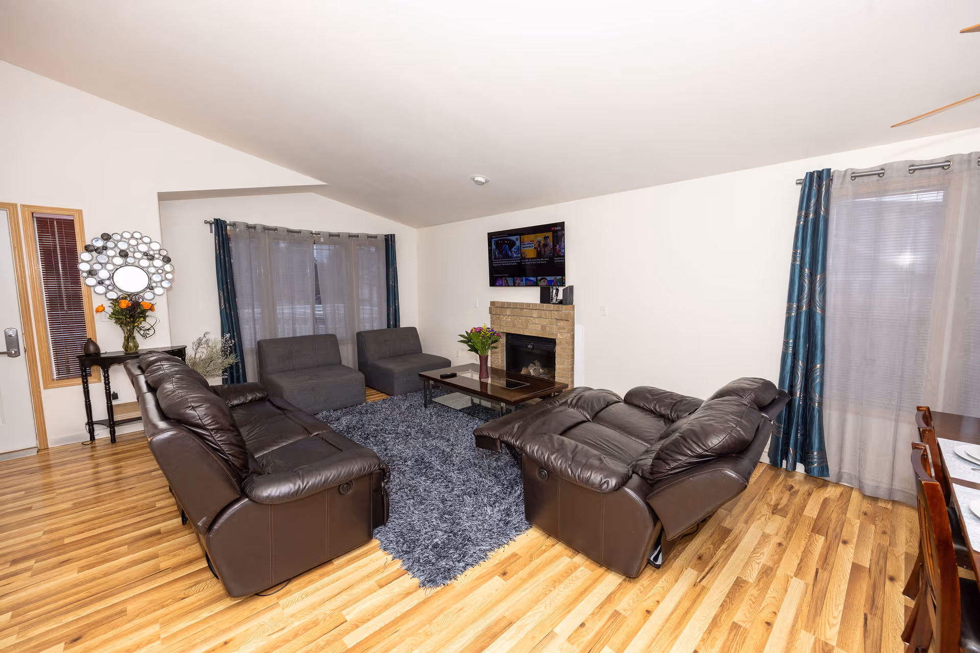 A cozy living room with two dark brown leather recliners and two gray armchairs arranged around a glass coffee table on a gray shag rug. There is a fireplace with a mounted flat-screen TV above it. The room has wooden flooring, white walls, and windows with blue curtains and sheer white drapes. A small table with a decorative mirror and a vase of flowers is near the door.