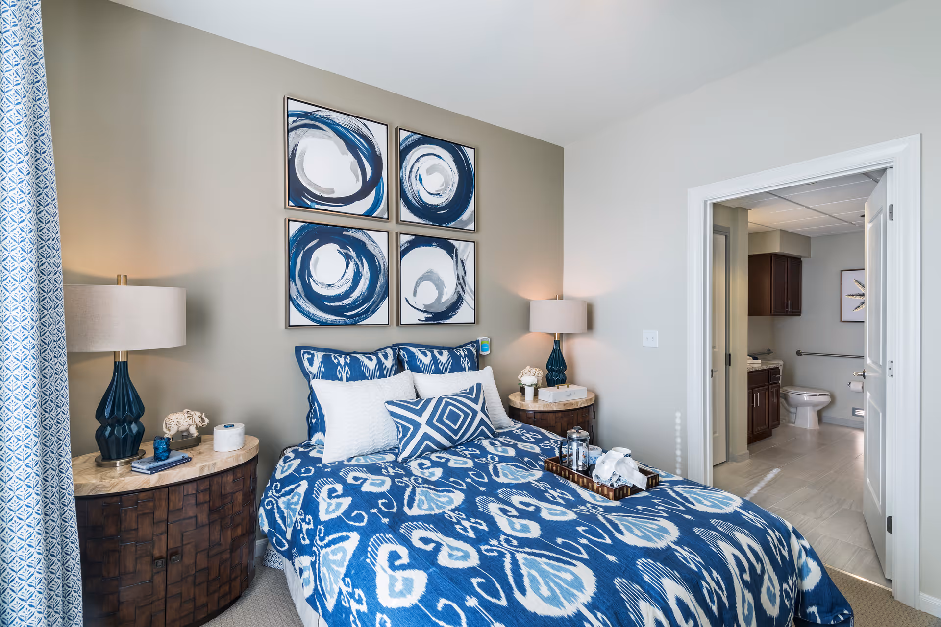 A cozy bedroom with a bed covered in blue and white patterned bedding, two matching bedside tables with lamps, and four abstract blue and white paintings on the wall above the bed. An open door reveals an adjacent bathroom with a toilet, sink, and wooden cabinets.