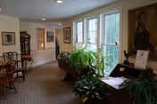 A well-lit interior hallway or common area with large windows on the right side letting in natural light. The space features several plants, framed artwork on the walls, and antique-style furniture including a wooden desk and chairs. The carpet has a subtle pattern, and the overall atmosphere is cozy and inviting.