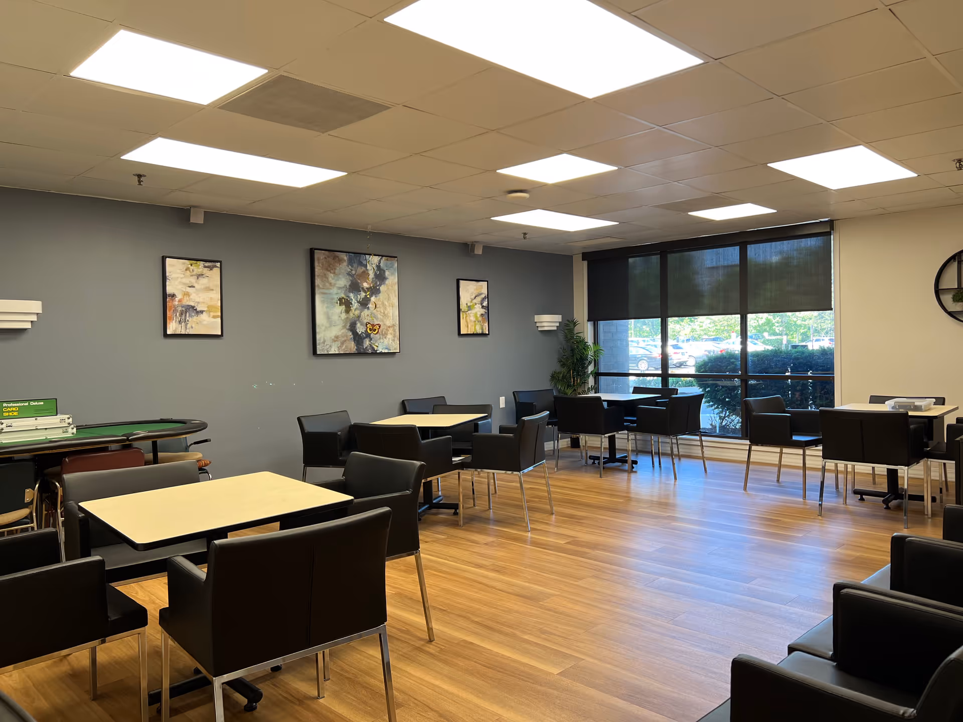 A well-lit room with several tables and black chairs arranged around them, wooden flooring, large windows with black roller shades partially down, and abstract paintings on a gray wall. There is a plant in the corner and a card table with a green felt surface on the left side.