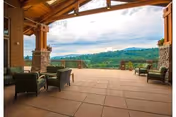 Covered outdoor patio area with cushioned chairs and a table, overlooking a scenic view of green hills and a partly cloudy sky.
