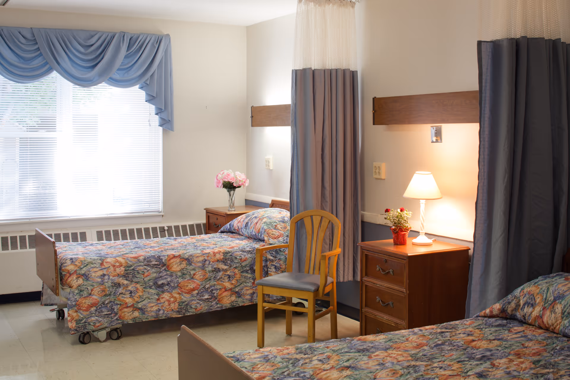 A shared bedroom in a senior living facility with two single beds covered in floral patterned bedspreads. Each bed has a wooden headboard and is separated by a wooden nightstand with a lamp and a small flower pot. A wooden chair is placed between the beds. The room has a large window with blue drapes and blinds, and privacy curtains hanging from the ceiling around each bed.