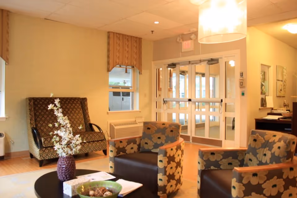 A cozy seating area in a senior living facility with two floral-patterned armchairs and a patterned loveseat. A round black coffee table with a vase of white flowers and a green bowl is in the center. The background shows glass double doors leading outside and a reception desk area to the right.