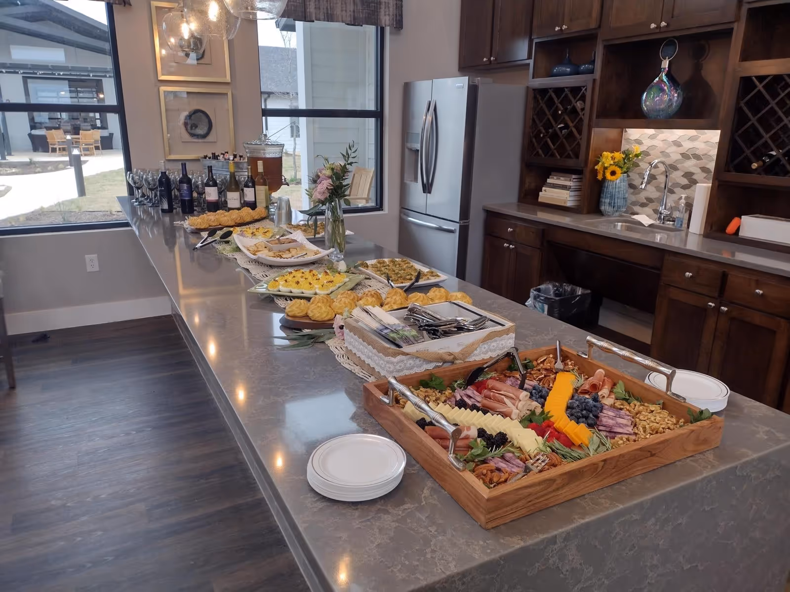 A modern kitchen area with a long marble countertop displaying an assortment of food including a charcuterie board with cheese, meats, nuts, and fruits, deviled eggs, pastries, and bottles of wine. The kitchen has dark wooden cabinets, a stainless steel refrigerator, a sink with a vase of sunflowers, and large windows showing an outdoor seating area.