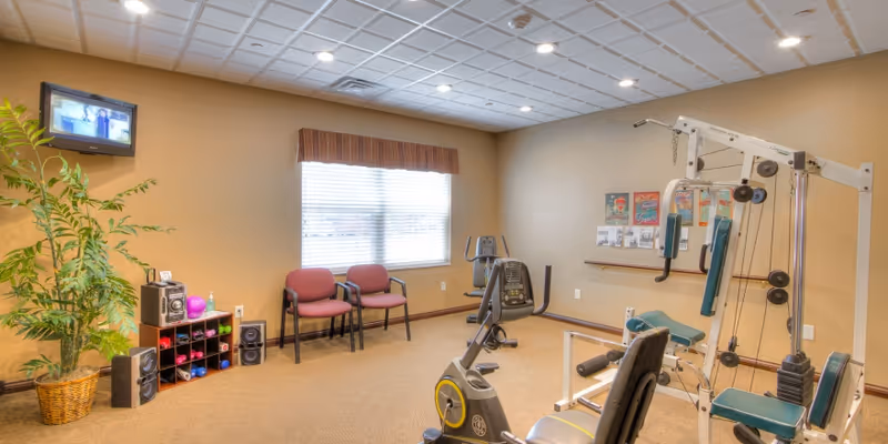A small fitness room with exercise equipment including a stationary bike, weight machine, and an elliptical trainer. The room has beige walls, a window with blinds and a valance, two red chairs, a potted plant, a small shelf with dumbbells, and a wall-mounted TV.