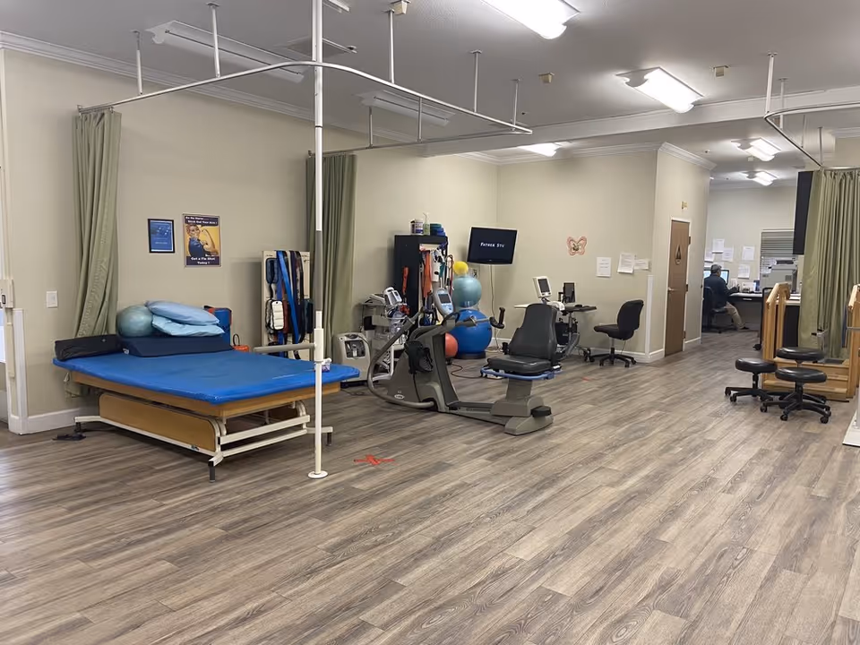 A rehabilitation or physical therapy room with a blue therapy bed, exercise equipment including a recumbent bike, therapy balls, and various medical devices. There are chairs, a desk with a computer, and a person working at the desk in the background. The room has wood-patterned flooring and light-colored walls with some posters and papers pinned up.