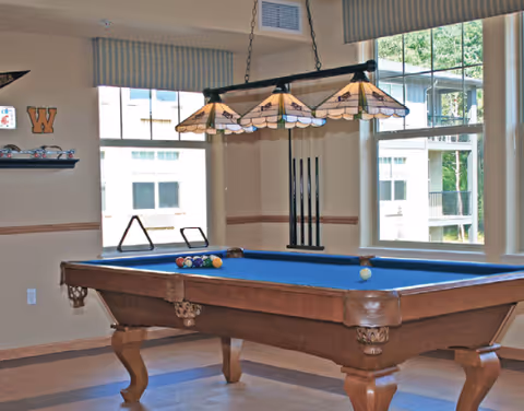 A pool table with blue felt under a stained-glass hanging light in a bright interior room with windows and a cue rack.