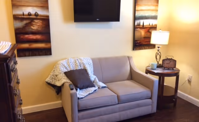 Cozy sitting area featuring a loveseat with a throw and pillow, a round side table with a lamp and clock, a wall-mounted TV, and framed artwork.