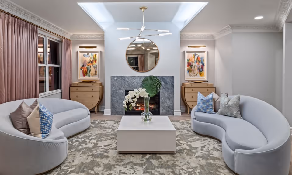 A modern living room with two curved light gray sofas facing each other across a square white coffee table with a glass vase holding white flowers and a green leaf. The room features a fireplace with a gray stone surround, a round mirror above it, and two wooden dressers on either side with colorful abstract paintings above them. Pink curtains cover a window on the left side, and a contemporary chandelier hangs from the ceiling.