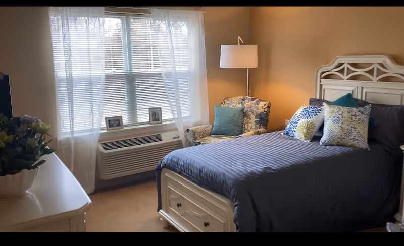 A cozy bedroom with a neatly made bed featuring a dark blue comforter and multiple decorative pillows. Next to the bed is a patterned armchair with a teal cushion and a tall floor lamp. A window with sheer white curtains and blinds lets in natural light. A white dresser with a potted plant and framed photos is visible in the foreground.