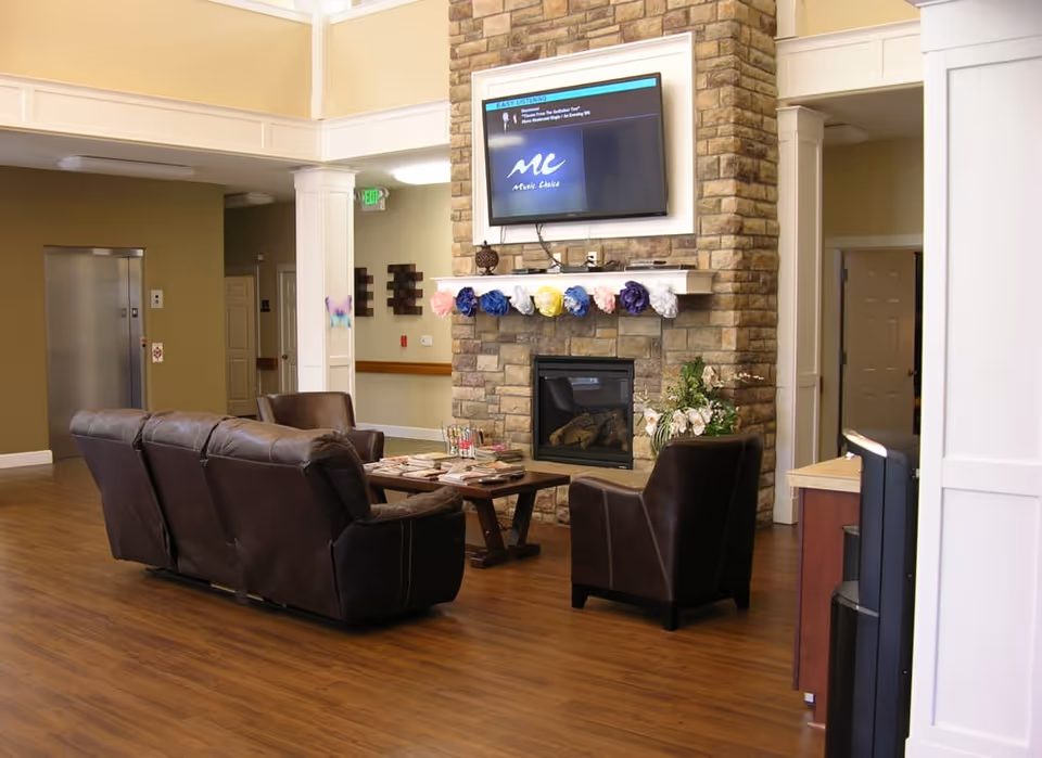 A cozy common area with brown leather sofas and chairs arranged around a wooden coffee table. A stone fireplace with a mantel decorated with colorful paper flowers is centered on the wall, above which a flat-screen TV is mounted. The room has wood flooring and neutral-colored walls, with an elevator visible in the background.