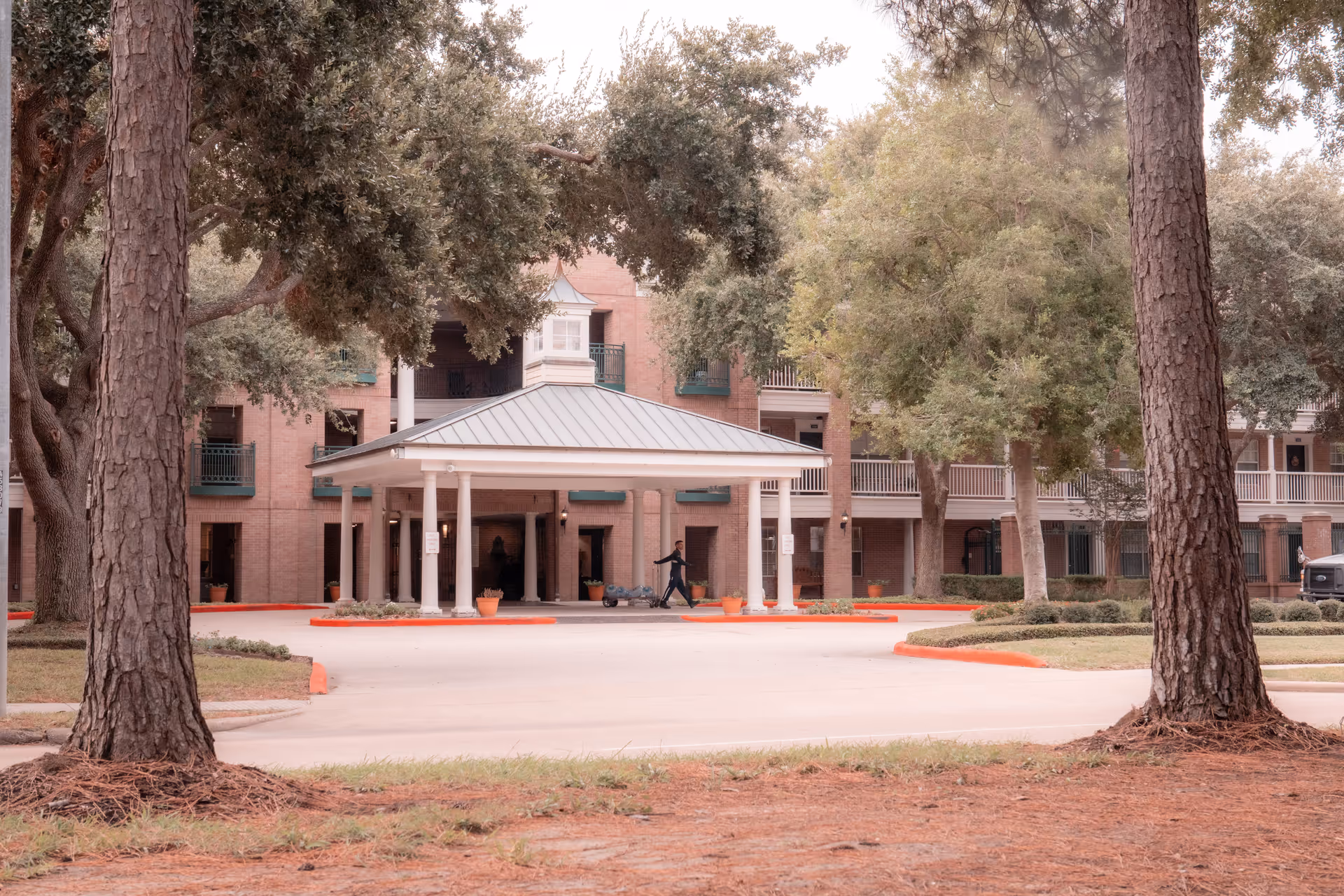 Front entrance of a brick senior living building with a covered porte-cochère framed by large trees.