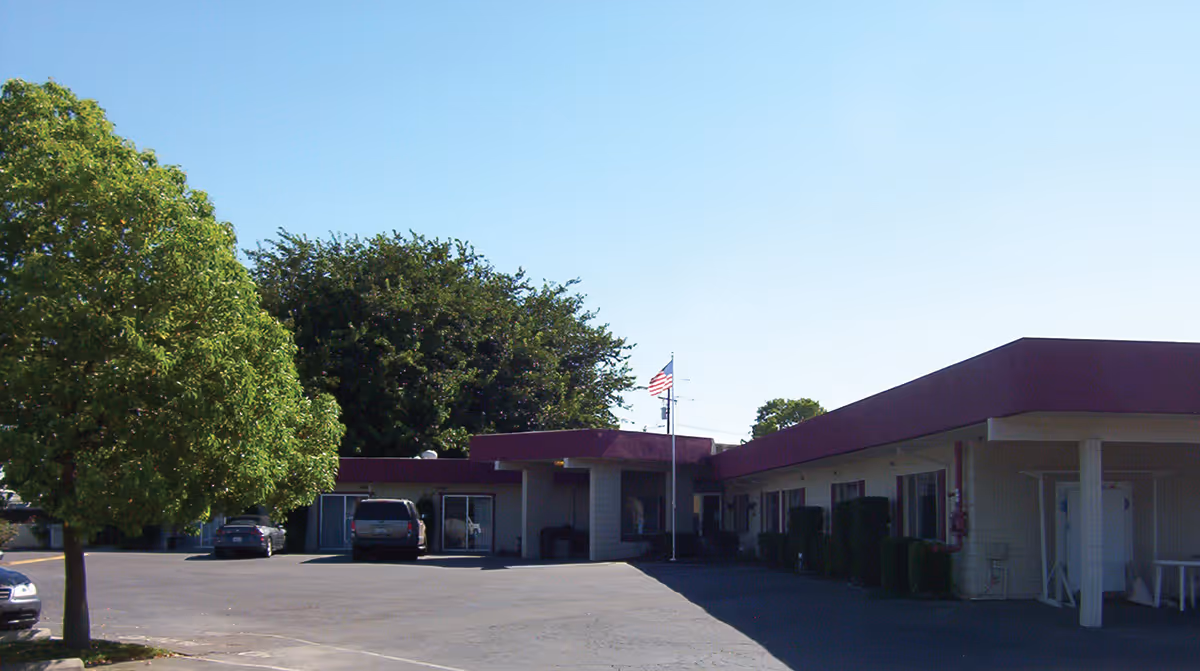 Exterior view of a single-story care facility building with a maroon roof trim, several windows, and an American flag on a flagpole in front. There are a few parked cars and green trees around the parking lot under a clear blue sky.