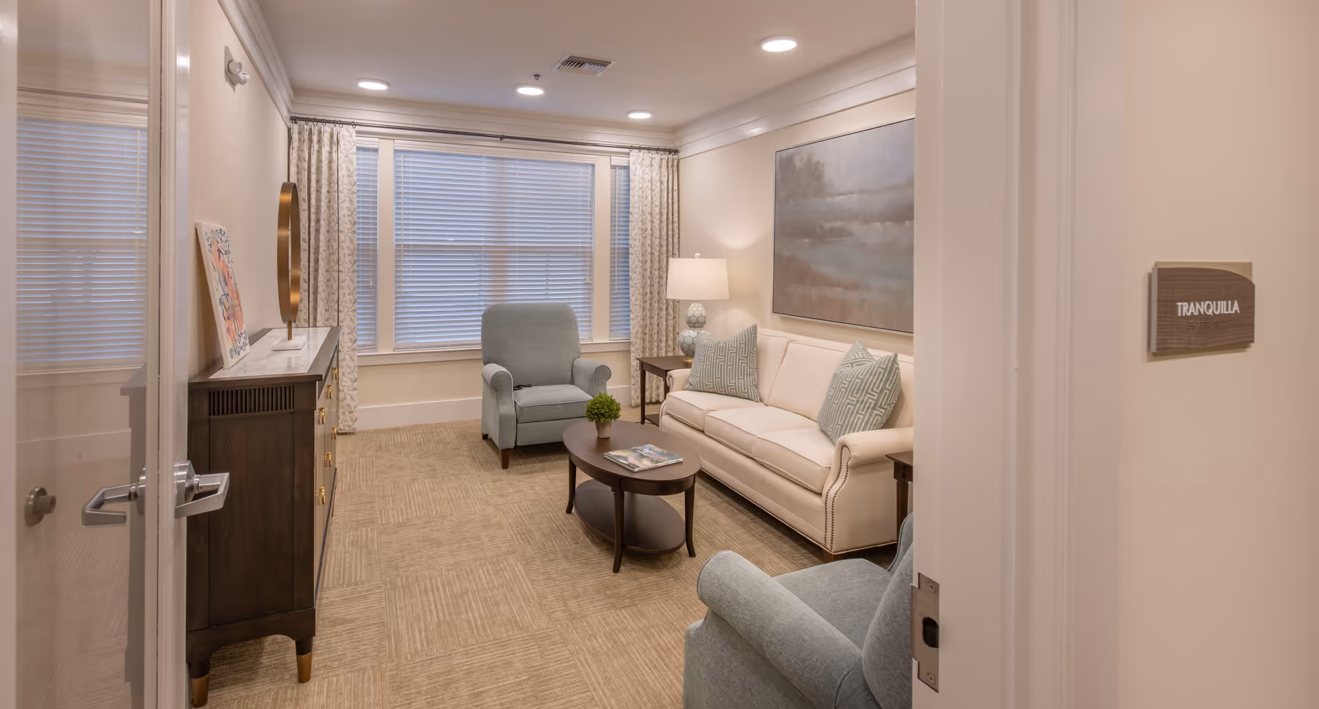 A cozy living room with a beige sofa, two blue armchairs, a round coffee table, sideboard and large window with blinds.