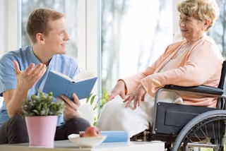 A young man in a blue shirt is sitting and holding a book while talking to an elderly woman in a wheelchair. They are indoors near a window with natural light, and a small potted plant and some fruit are on the table in front of them.