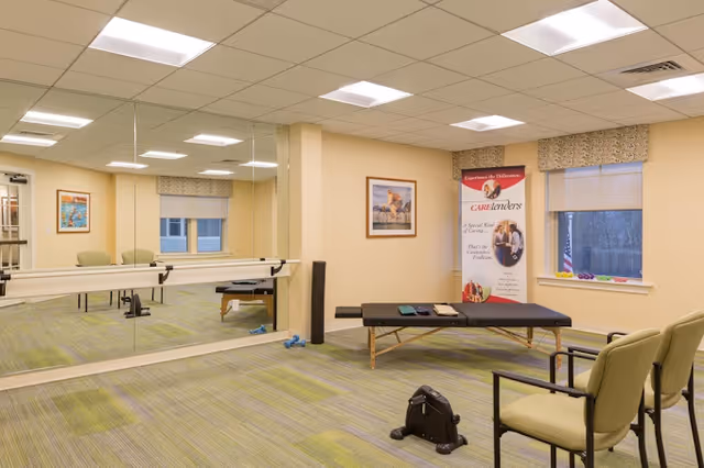 A bright interior therapy/exercise room with a mirrored wall and barre, a treatment table, chairs, and a promotional banner.