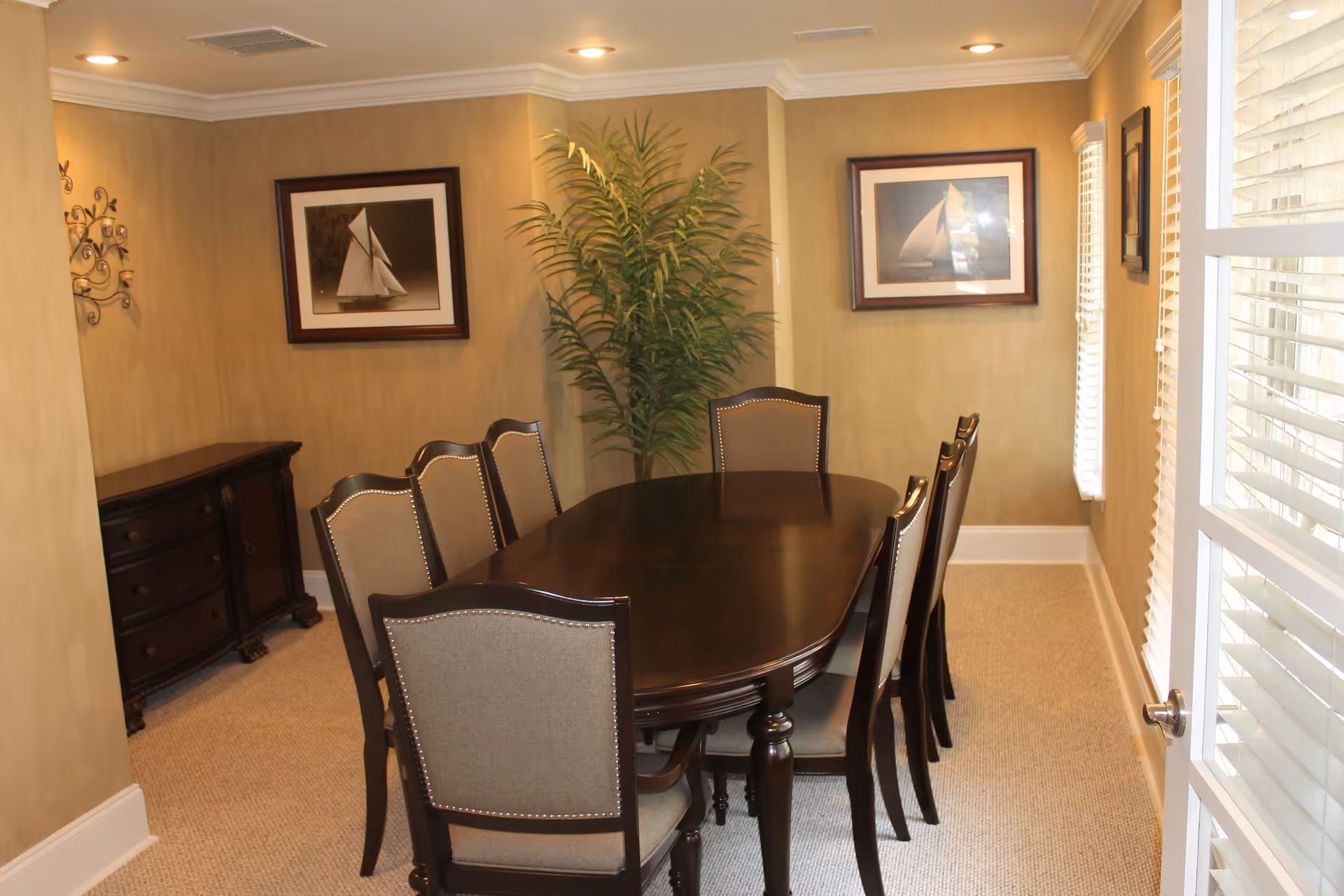 A dining room with a dark wooden table surrounded by eight upholstered chairs. The walls are beige with two framed pictures of sailboats. There is a tall green plant in the corner and a dark wooden sideboard against one wall. The room is lit by recessed ceiling lights and natural light coming through windows with white blinds.