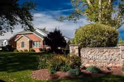 A stone entrance sign reading 'WOODLANDS' sits amid landscaped grounds in front of a brick residential building under a blue sky.