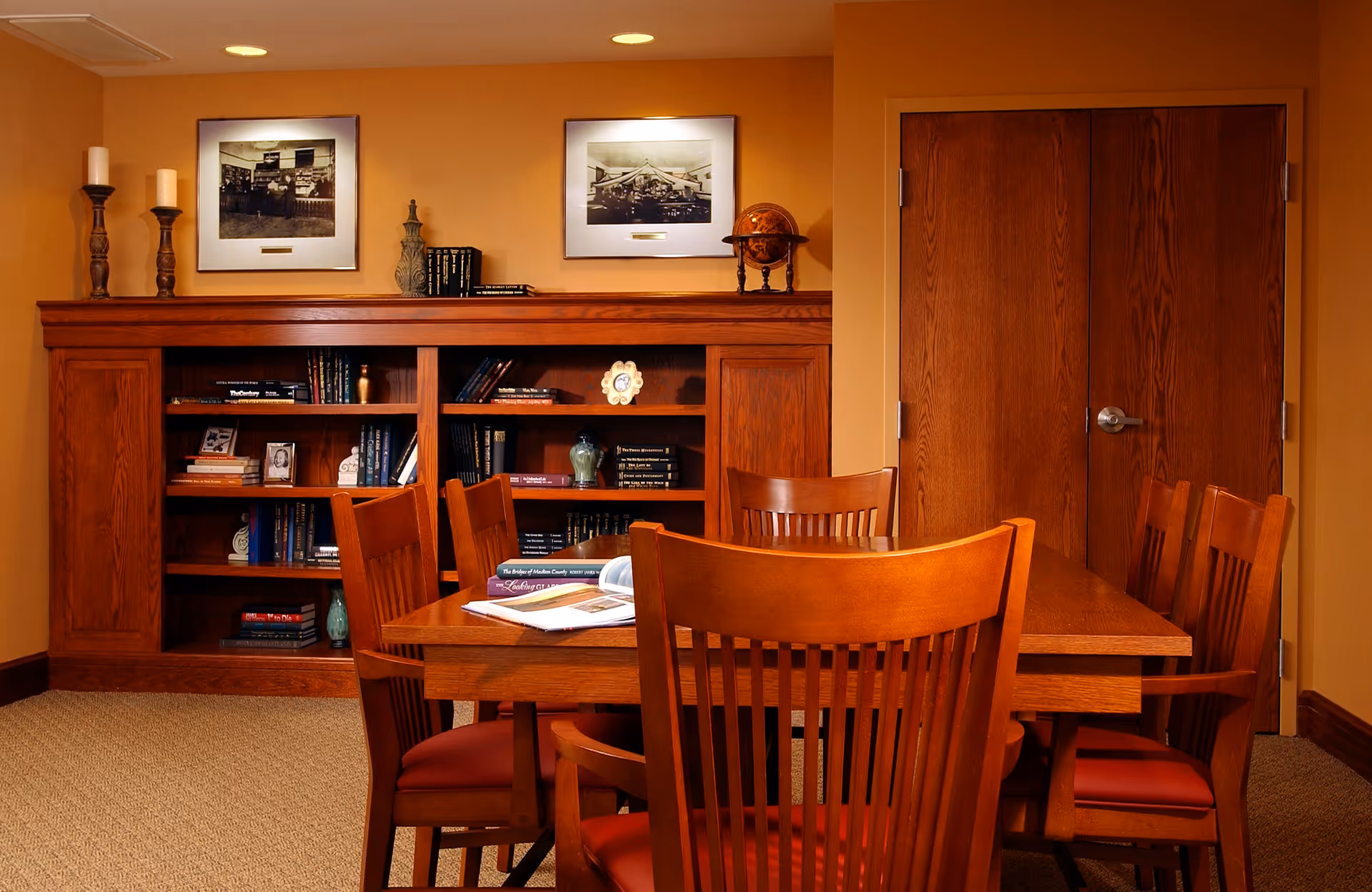 A cozy interior room with a wooden table surrounded by six wooden chairs with red cushions. Behind the table is a wooden bookshelf filled with books and decorative items, including candles, a globe, and framed black and white photographs on the wall. The walls are painted a warm beige color, and there is a closed wooden double door to the right.