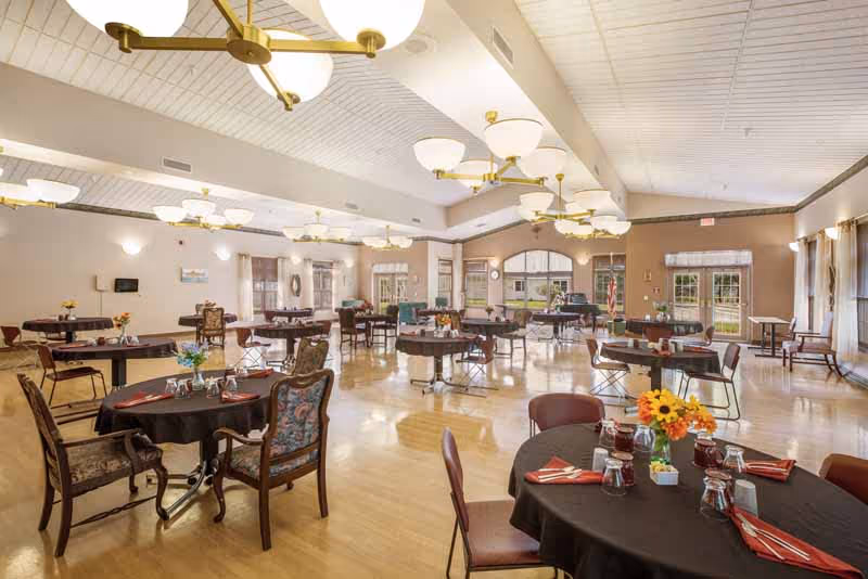 Spacious dining room with multiple round tables covered with black tablecloths, each set with glasses, napkins, and small flower arrangements. The room has large windows letting in natural light, wooden flooring, and several ceiling light fixtures with globe-shaped lamps.