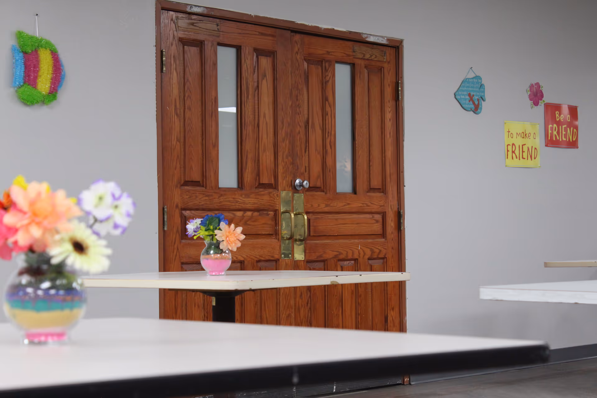Interior view of a room with wooden double doors in the background. The room has white tables with small vases of colorful artificial flowers placed on them. The walls are decorated with colorful signs and wall hangings, including messages like 'to make a FRIEND' and 'Be a FRIEND'.