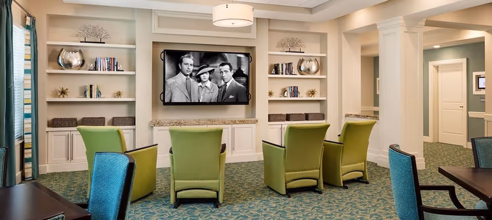 Community lounge with green chairs facing a wall-mounted TV and built-in shelving.