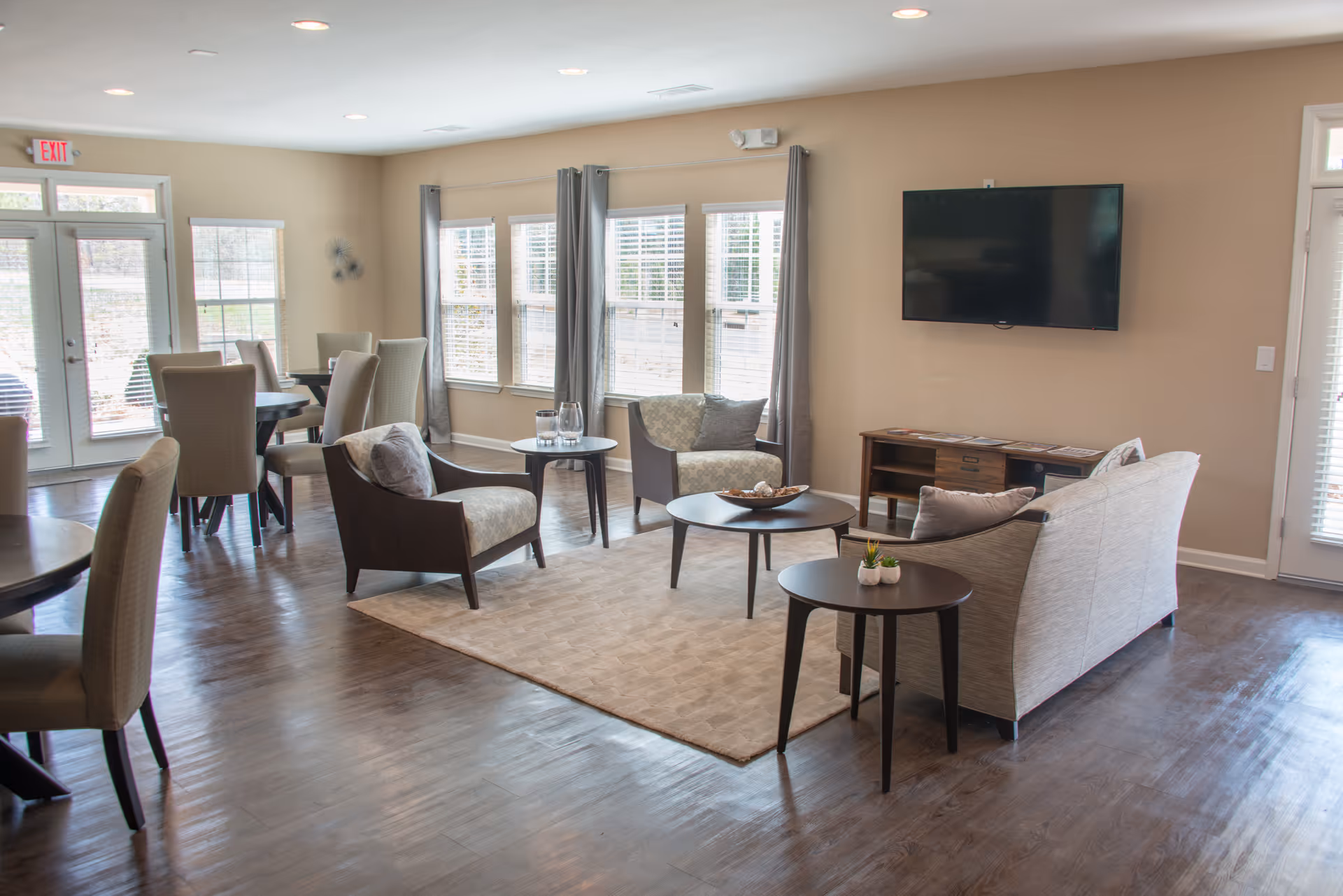 Bright communal living area with sofas, armchairs, round coffee tables, dining tables, and a wall-mounted TV.