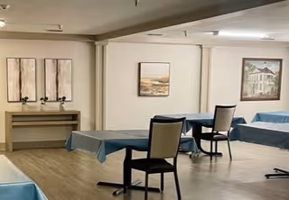 Interior view of a common area with several tables covered with blue tablecloths and chairs arranged around them. The room has light-colored walls adorned with framed artwork and a small console table against one wall. The floor is wooden, and the space appears clean and well-lit.