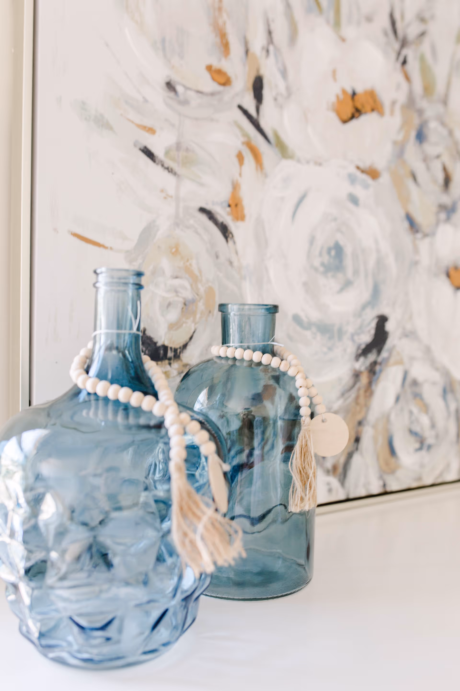 Two decorative blue glass bottles adorned with white bead garlands and tassels placed on a white surface in front of a floral abstract painting with white, beige, and black tones.