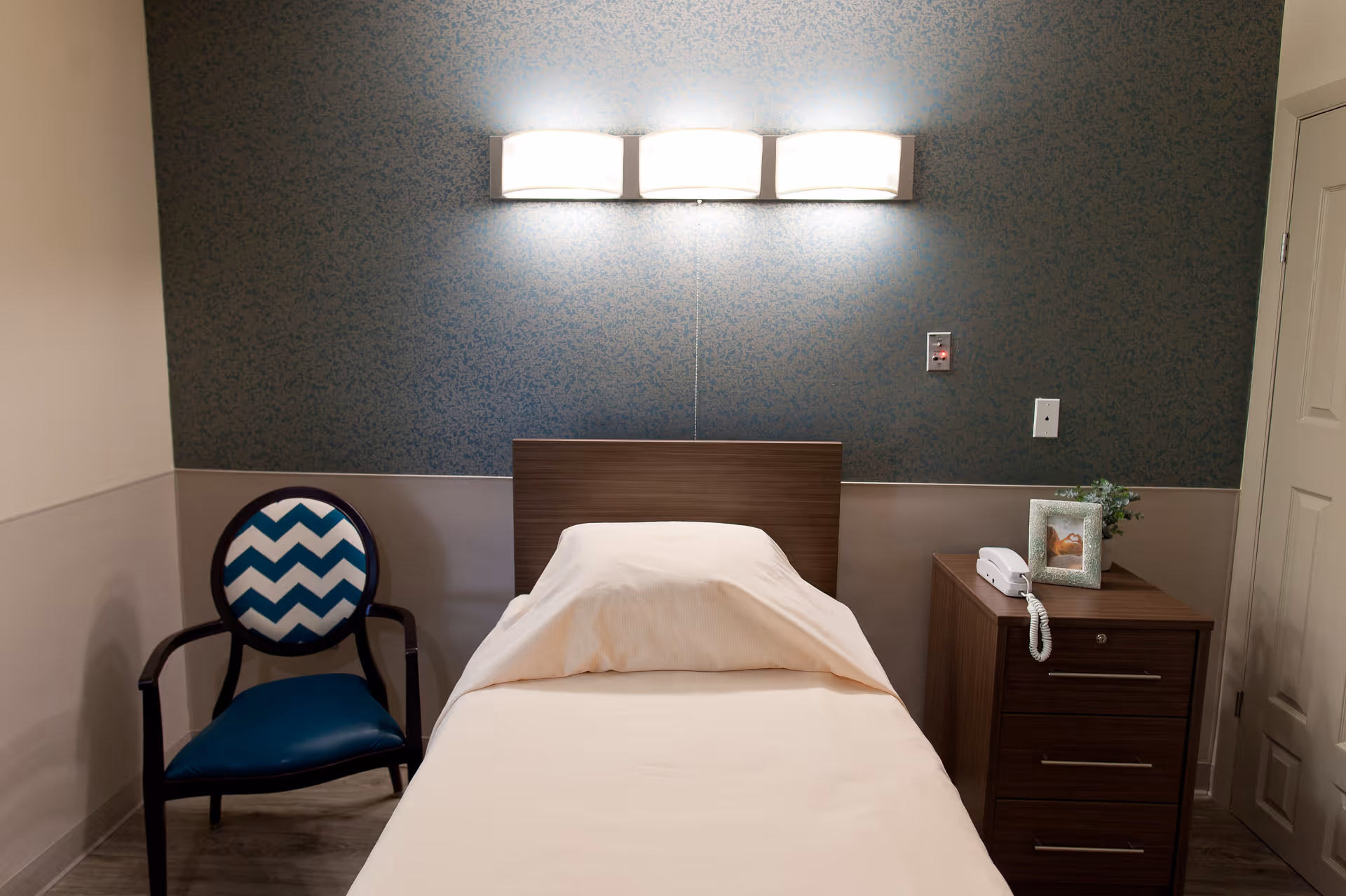 A small resident bedroom with a single bed, wooden headboard, wall light, bedside table with a phone and framed photo, and a patterned chair.