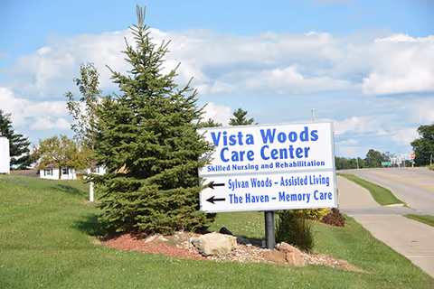 A roadside sign for Vista Woods Care Center, a skilled nursing and rehabilitation facility, with directional arrows pointing to Sylvan Woods - Assisted Living and The Haven - Memory Care. The sign is placed on a grassy area with a small evergreen tree and rocks nearby, under a partly cloudy blue sky.