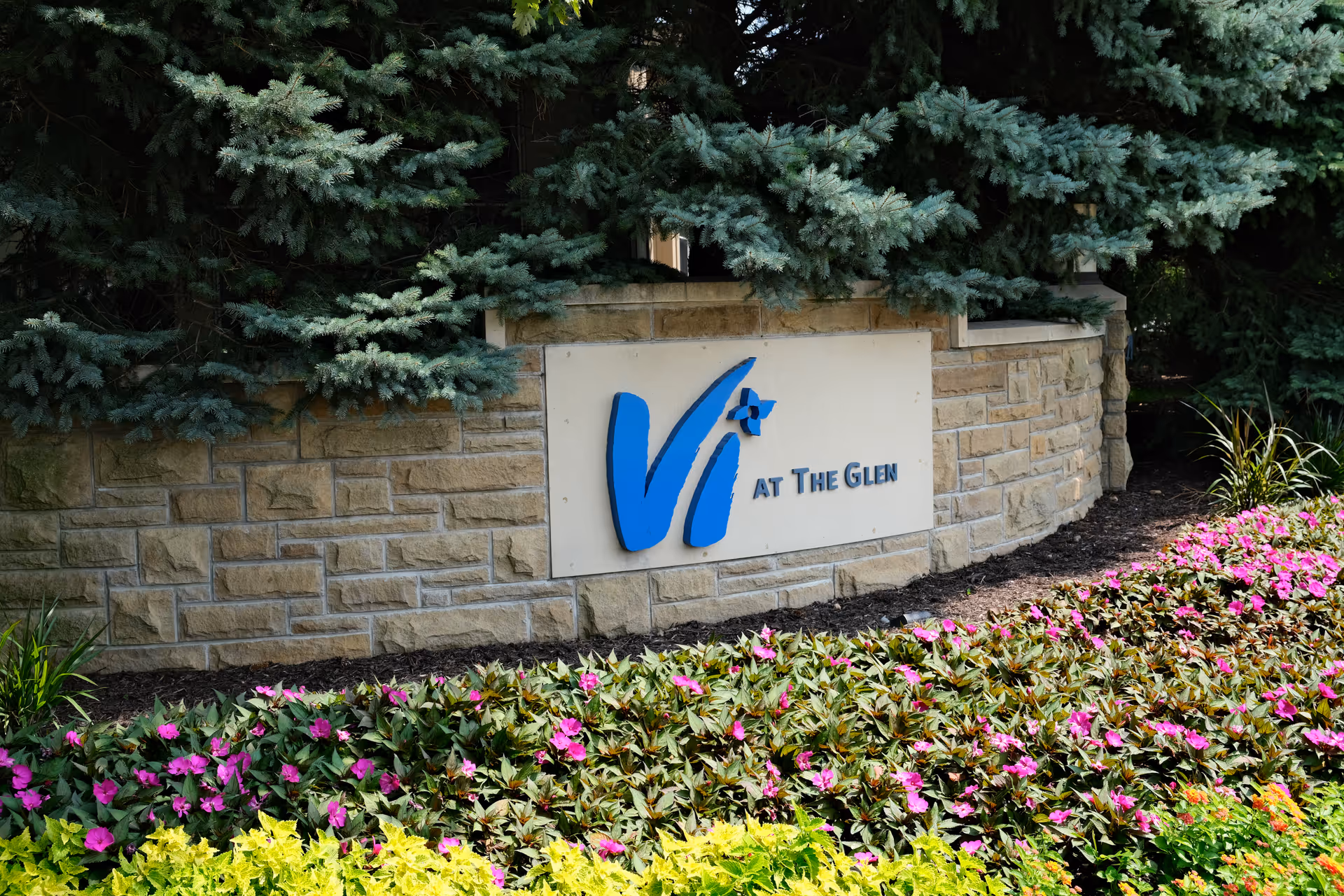 Stone entrance sign reading 'Vi at The Glen' surrounded by evergreen trees and colorful flower beds.